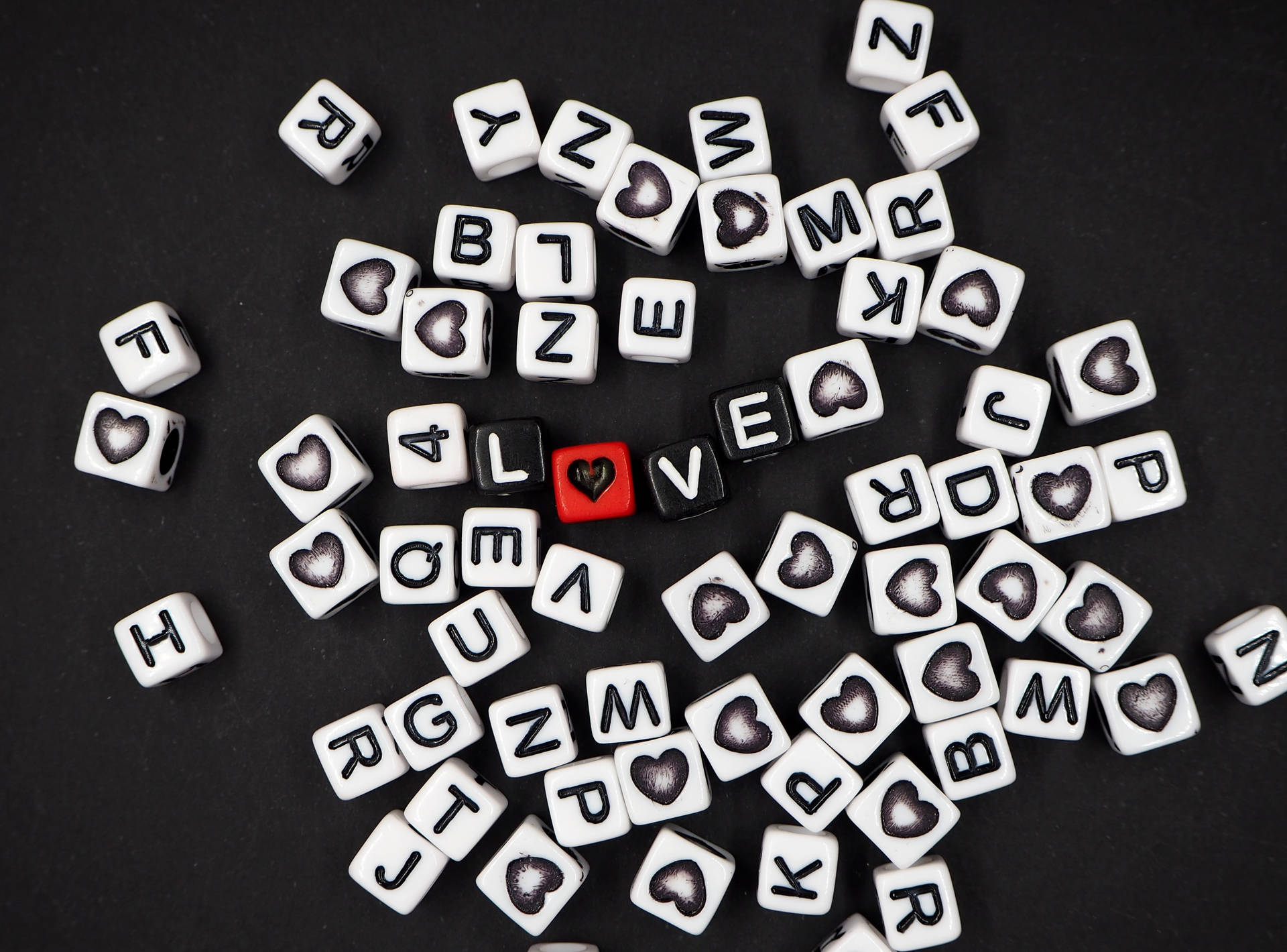 黒いサイコロが「love」という単語を形作り、「O」の代わりにハートがあしらわれ、白いサイコロの文字が黒い背景に散らばっています。ハートの日のために。