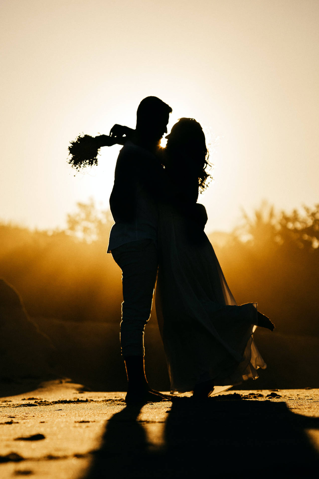 花束を持った花嫁と抱き合う結婚式のカップルのシルエット、夕日と黄橙色の空を背景に。