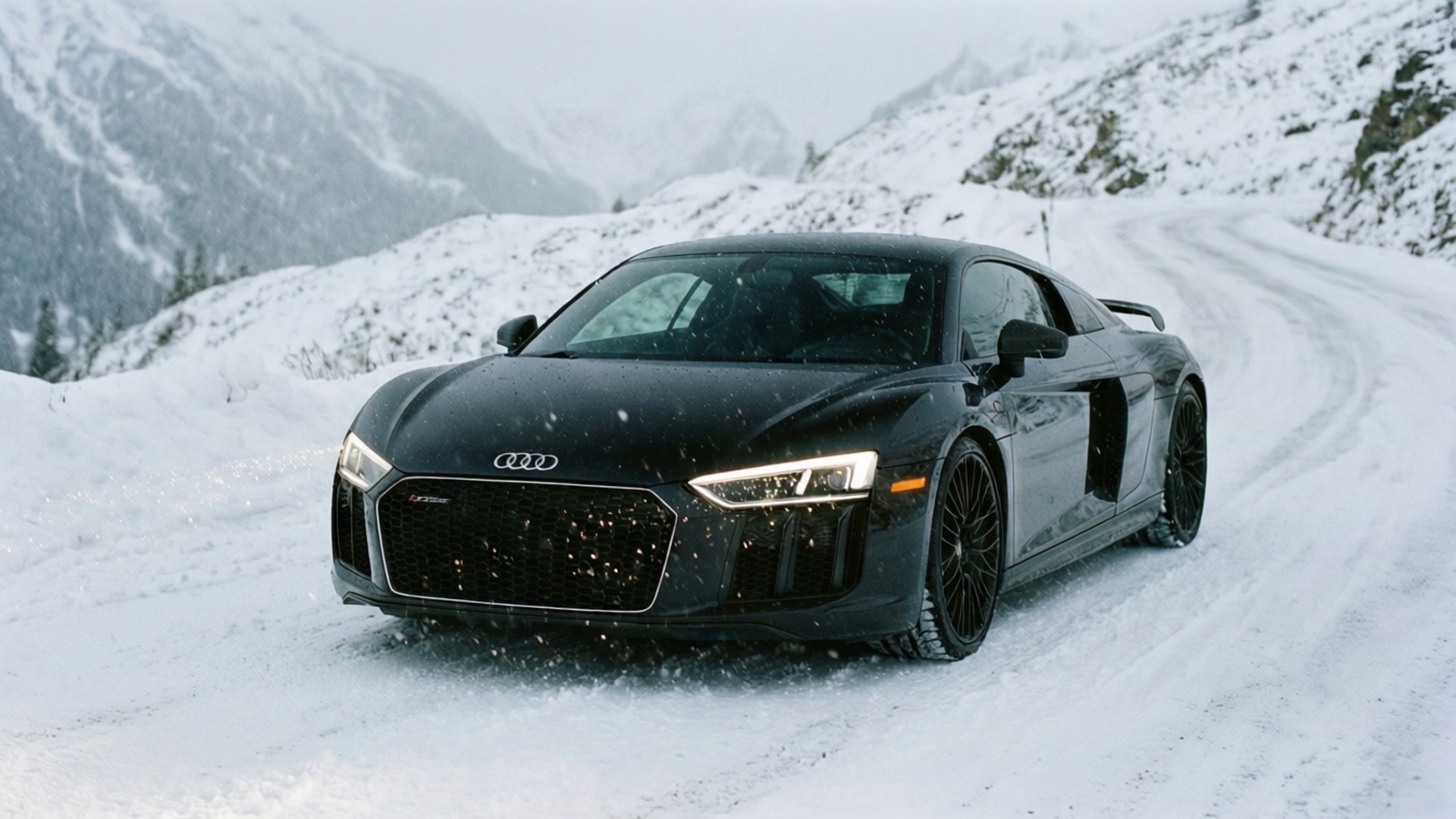 雪が降るアルプスの山道を走る黒いアウディR8。