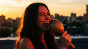 ビボがマラカスを手に、屋上で夕日を背景に心のこもった歌を披露している。